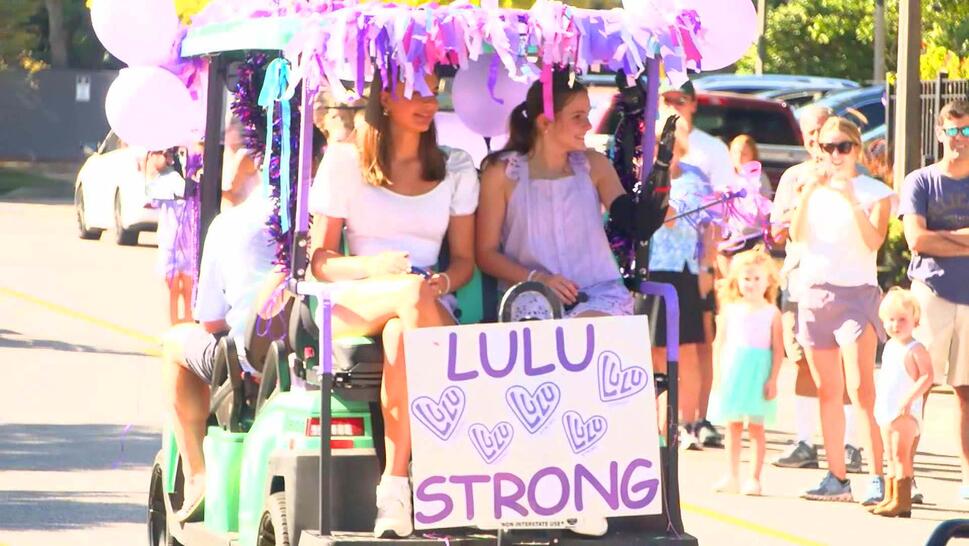 Golf cart driving down a street with a girl waving to the crowds lining the street. Poster on golf card reads: Lulu Strong